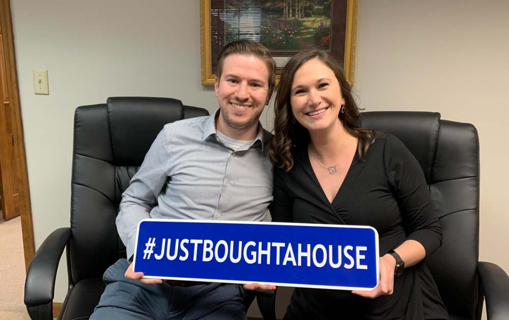A man and woman sit in chars, facing towards the camera, smiling. They're holding a sign that reads: #justboughtahouse.
