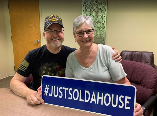 Happy couple holding a sign that says hashtag just sold a house while smiling and sitting at a table.