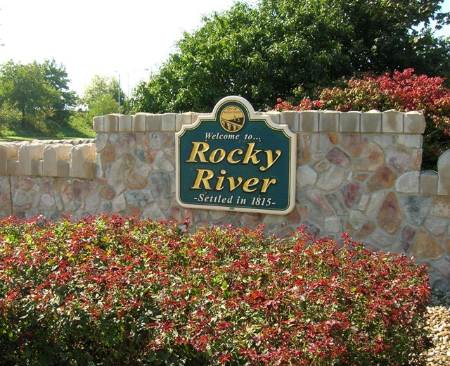 Green and gold city sign on a landscaping stone wall reading "Welcome to Rocky River settled in 1815" with a flower bed below located near homes for sale in Rocky River Ohio