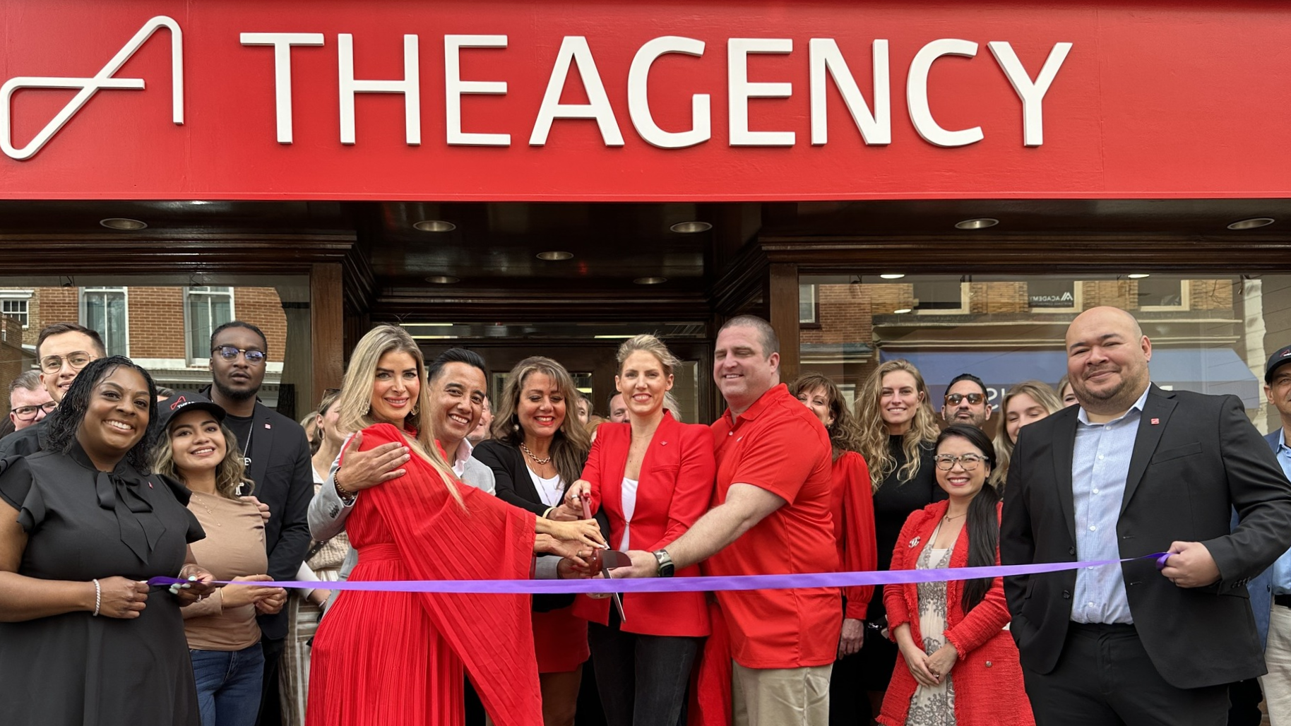 The 3 co-managing partners are holding a giant scissors open over a lavender ribbon. They are surrounded by loved ones, staff, and other regional teams from The Agency. The lavender ribbon is being held across the length of the crowd by 2 of The Agency Frederick's most skilled realtors; Joi Moses(left) and Antonio Meza(right)