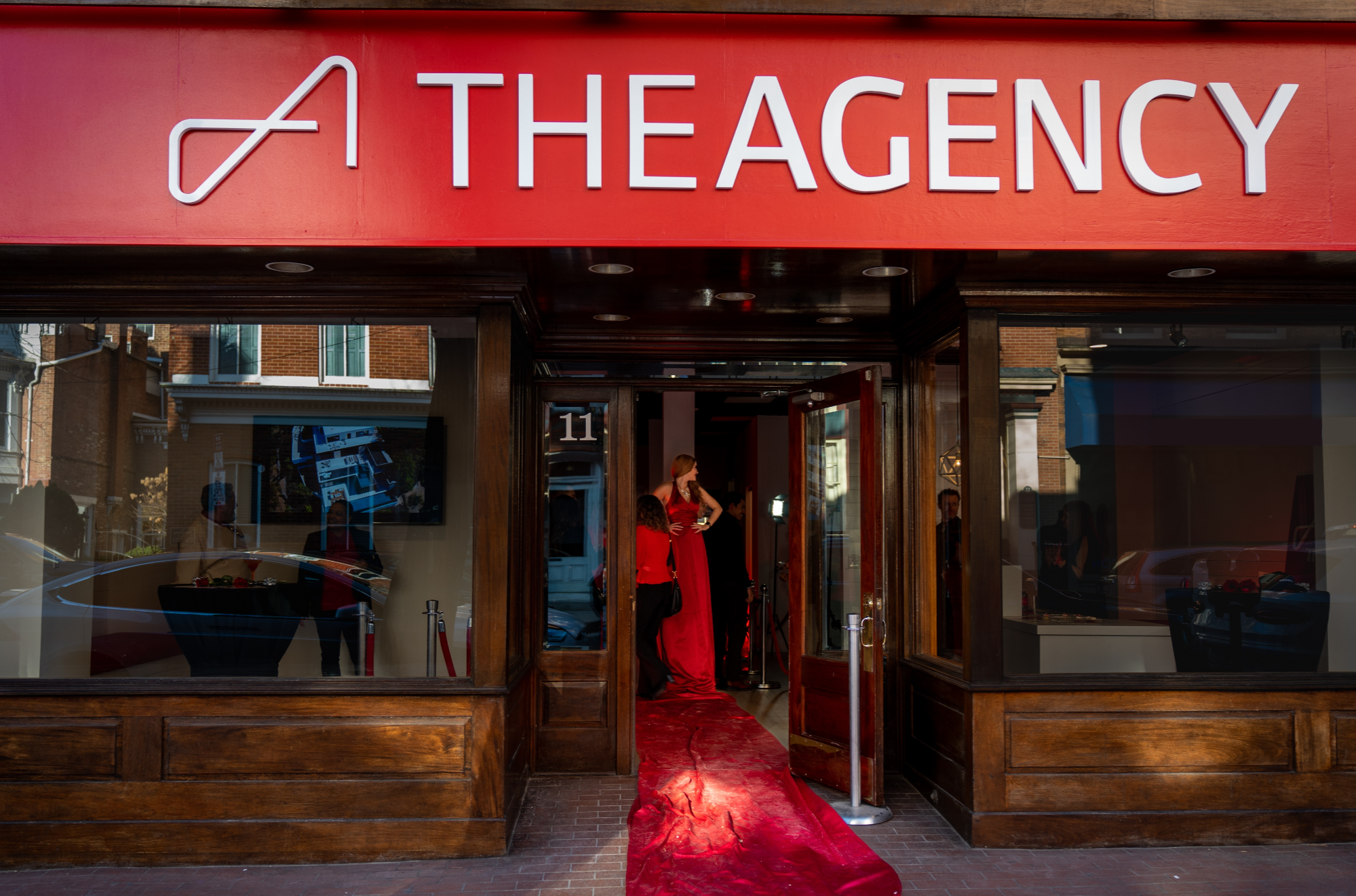 The Agency Frederick Facade. Through the open door a statuesque, RED haired woman is posing with her hands on her hips. Her evening gown is a vibrant RED and its train forms a posh RED carpet leading from inside the business to the redbrick sidewalk outside.