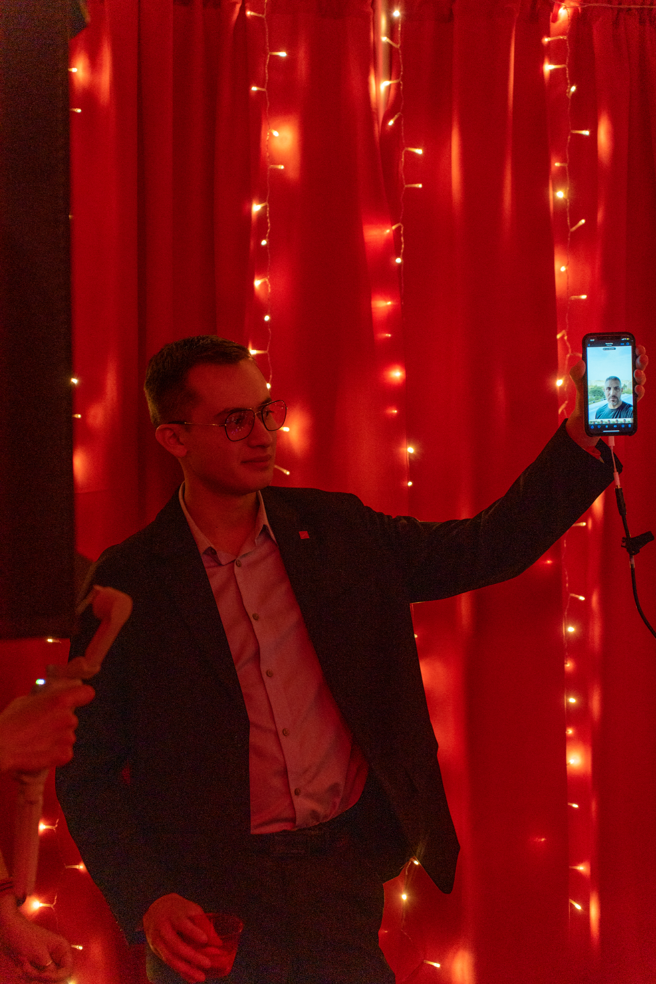 The Agency Frederick's Realtor Daniel Ramierez on FaceTime With The Agency CEO Mario Umansky . Daniel is dressed in a comfort-fit navy pants suit, and white collared shirt. He holds the glowing phone up to a completely RED room, so that The Agency CEO, Mario Umansky can see the grand affair and share his rousing words with the guests and co-managers.