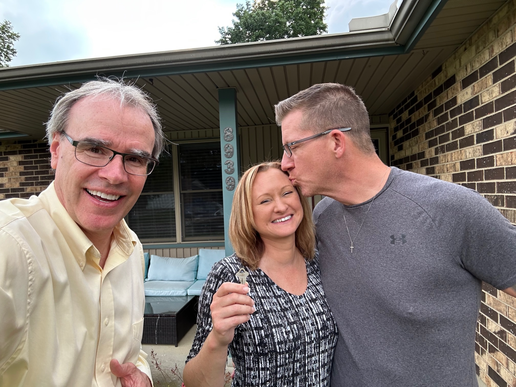 Two happy clients - a man and woman - stand next to David Campbell, Realtor - in front of their new house. The woman is holding a house key while the man is kissing her on the head.