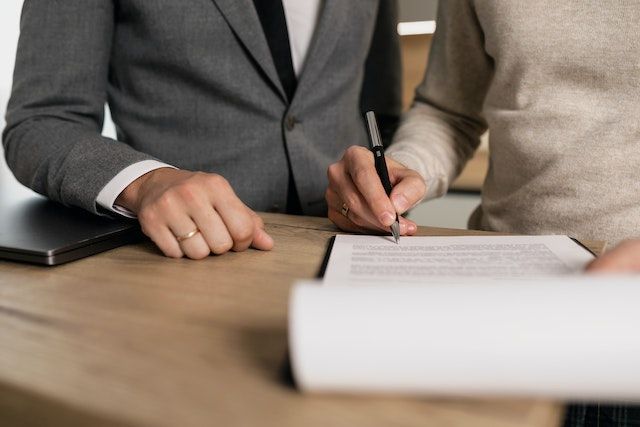 Person signing a home buying contract while standing next to their real estate agent