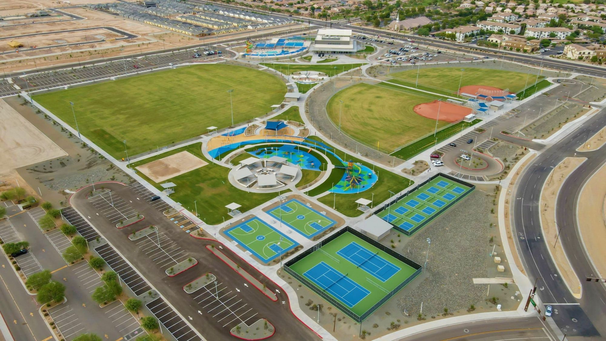 Aerial view of Goodyear Community Park, showcasing large open fields, sports courts, playgrounds, and recreational facilities. This expansive park in Goodyear, Arizona, offers a variety of amenities for residents, including basketball courts, tennis courts, splash pads, and baseball fields, making it a hub for outdoor activities and community gatherings.