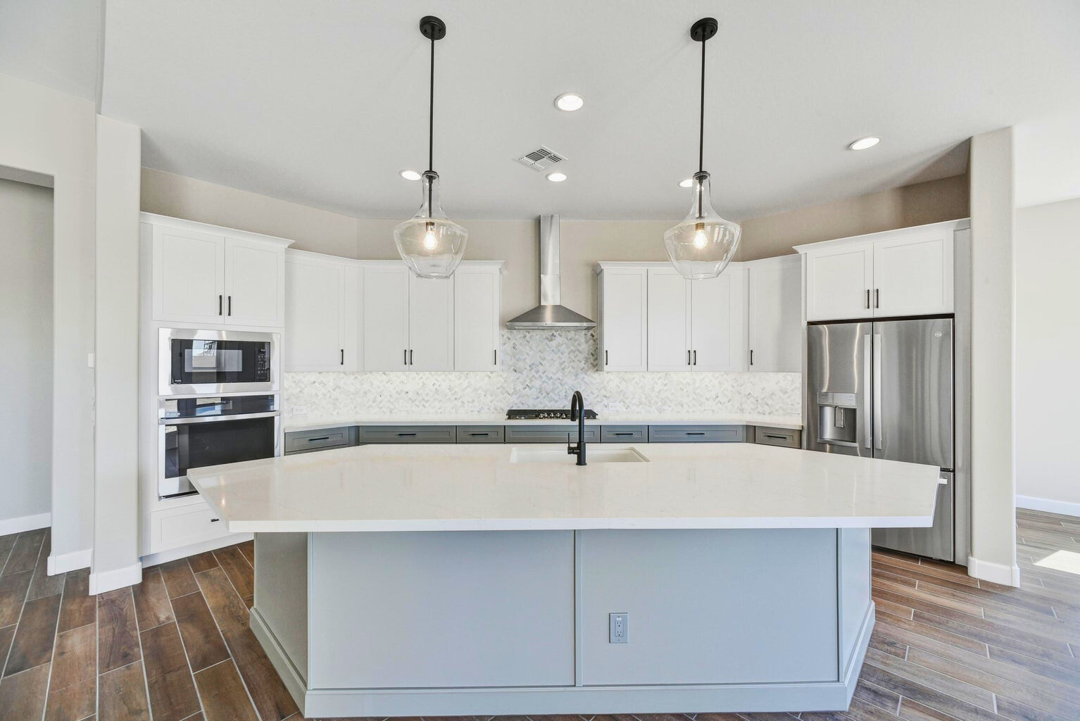 Stunning gourmet kitchen in a new construction home in Sentiero at Windrose, Waddell, AZ, featuring an oversized island with quartz countertops, two-tone cabinetry, stainless steel appliances, a gas cooktop, and modern pendant lighting. The elegant custom backsplash and spacious design make this kitchen ideal for entertaining and family gatherings.