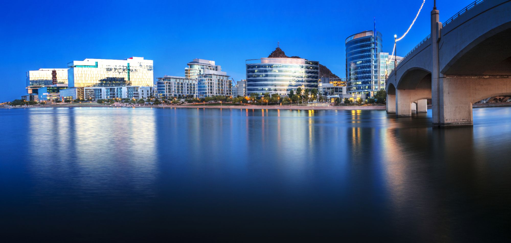 Scenic view of Tempe Town Lake at dusk with modern buildings and a bridge lit up, showcasing the vibrant atmosphere and exciting activities in Tempe, Arizona, including water sports, outdoor recreation, and lively urban experiences.