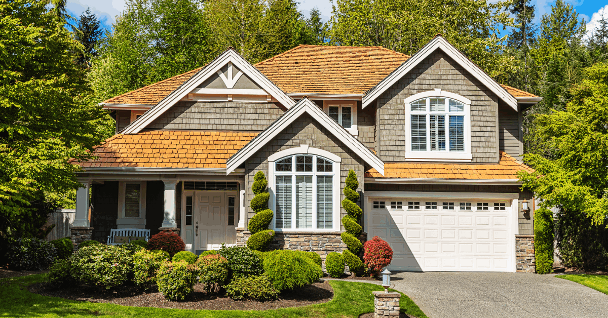 A simple but charming little house flanked by two taller houses. The house has a green lawn, a porch and a low iron fence.