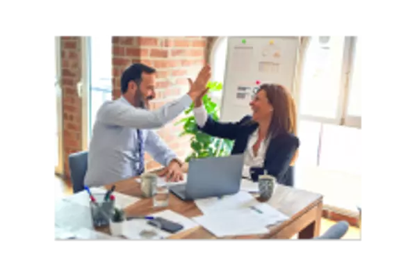 Two professionals high-fiving in a bright office setting, seated at a wooden table with a laptop, documents, and coffee cups.