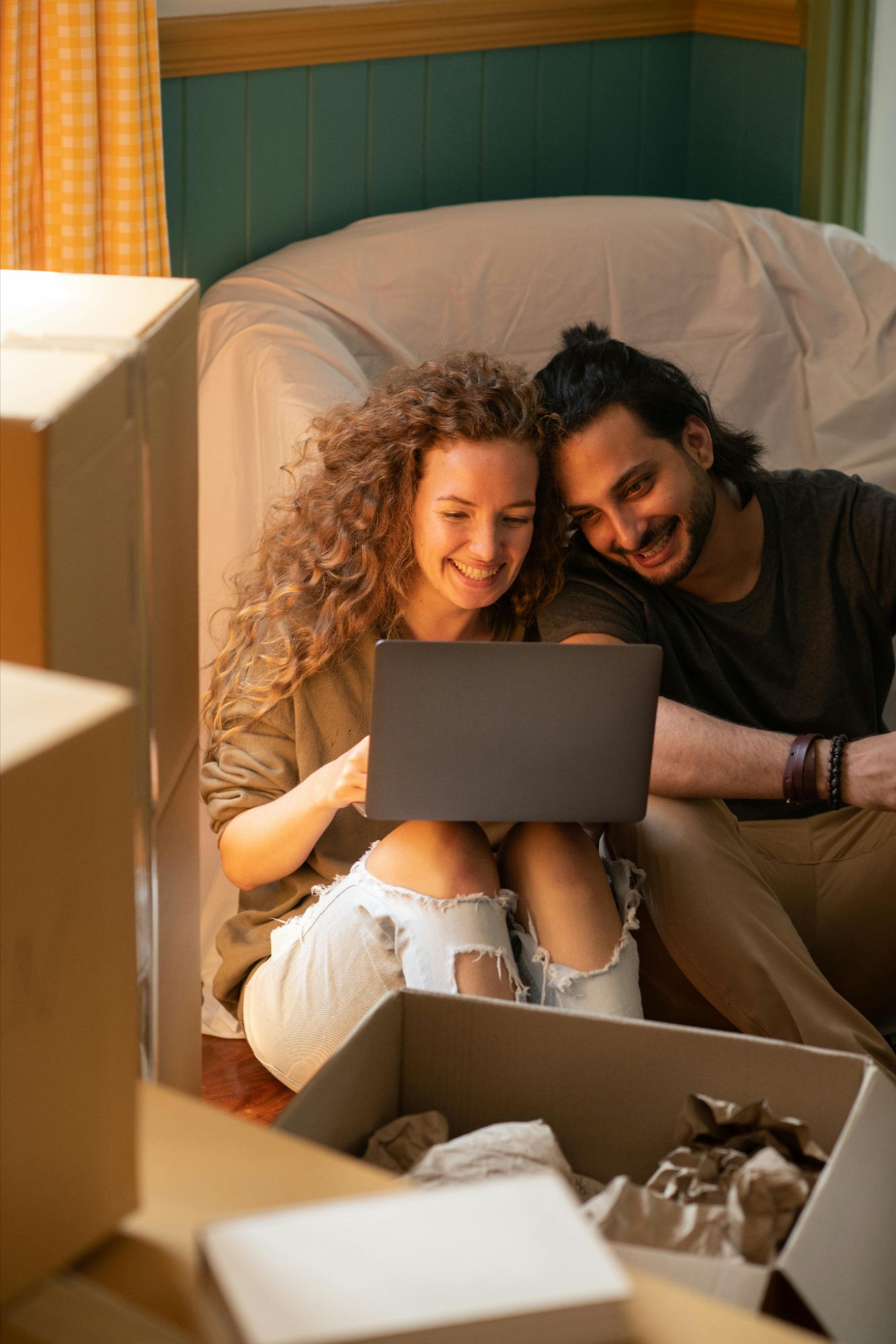 A couple sitting together in front of their computer, surrounded by moving boxes, unpacking items in their new home