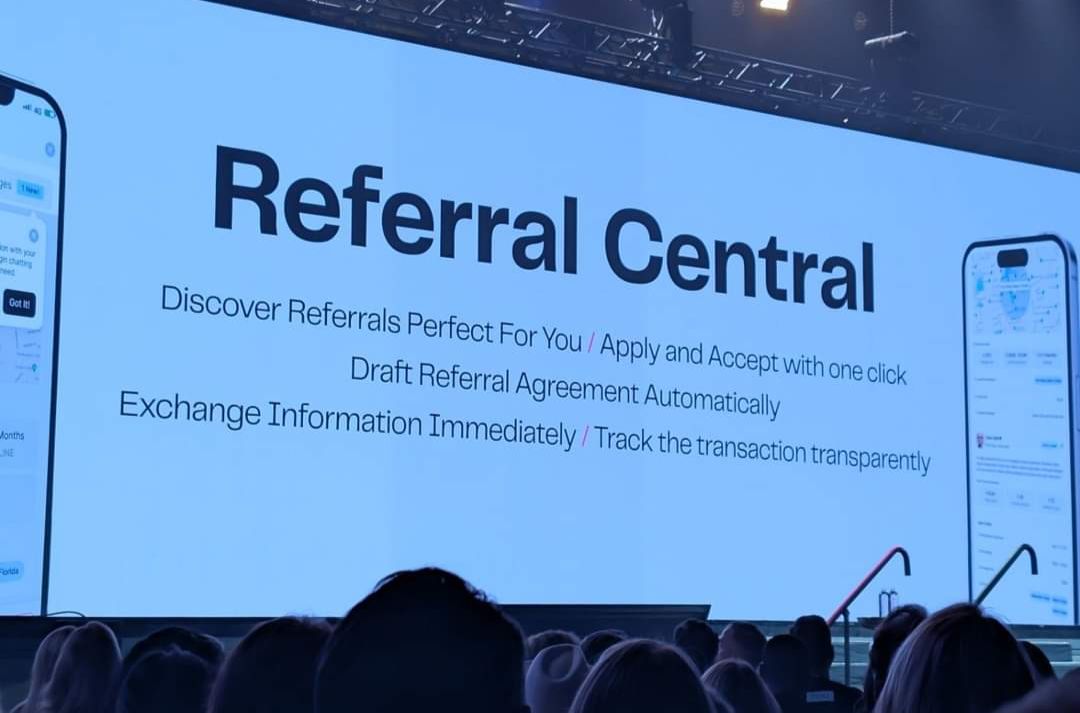 Real estate agents view a large screen presentation introducing 'Referral Central,' a new system designed to streamline referral processes. Key features include discovering ideal referrals, applying and accepting with a single click, automatic drafting of referral agreements, instant information exchange, and transparent transaction tracking.