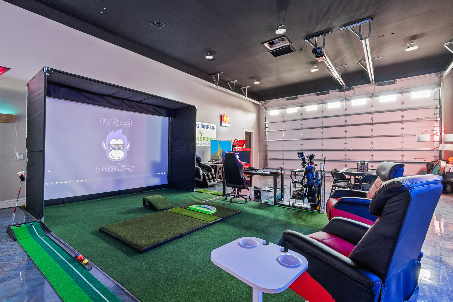 The image shows a modern indoor entertainment and golf simulation room set up in a large garage. The space features a golf simulator with a large screen and putting green, complete with golf clubs and equipment. In front of the screen, there are two reclining chairs with cup holders, creating a comfortable viewing area. Additional seating and a desk with computer equipment are visible to the side, along with various decorations and signage on the walls. The room has high ceilings, a polished floor, and large garage doors, making it a versatile recreational space.