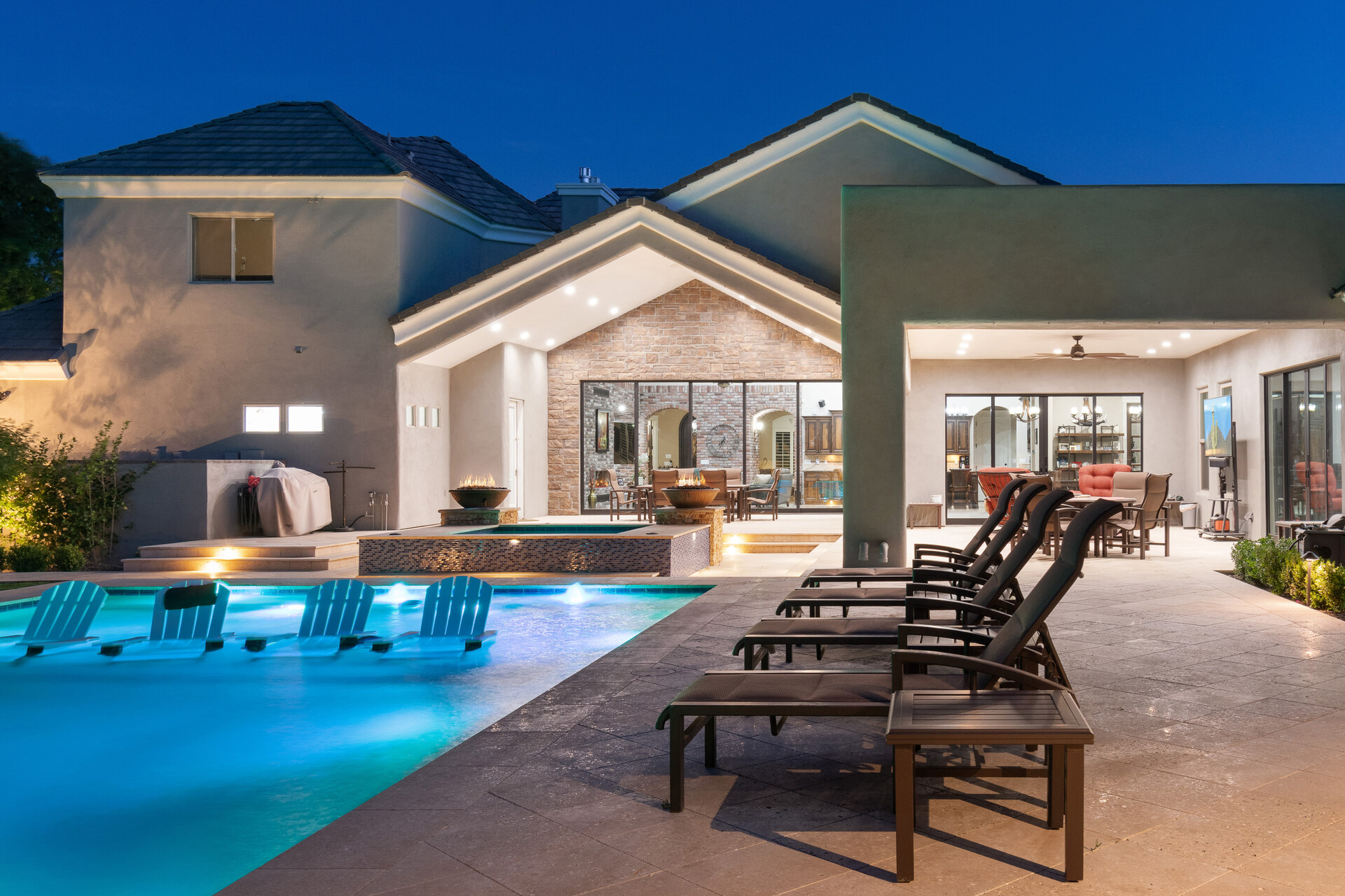 The image shows a modern backyard with a lit pool at night. Lounge chairs line the pool's edge, and the pool includes built-in seating. The house in the background features large glass doors, a covered patio with seating, and outdoor lighting that highlights the stone accents and architectural details. A grill and fire features add to the luxurious ambiance, creating a relaxing and inviting space.