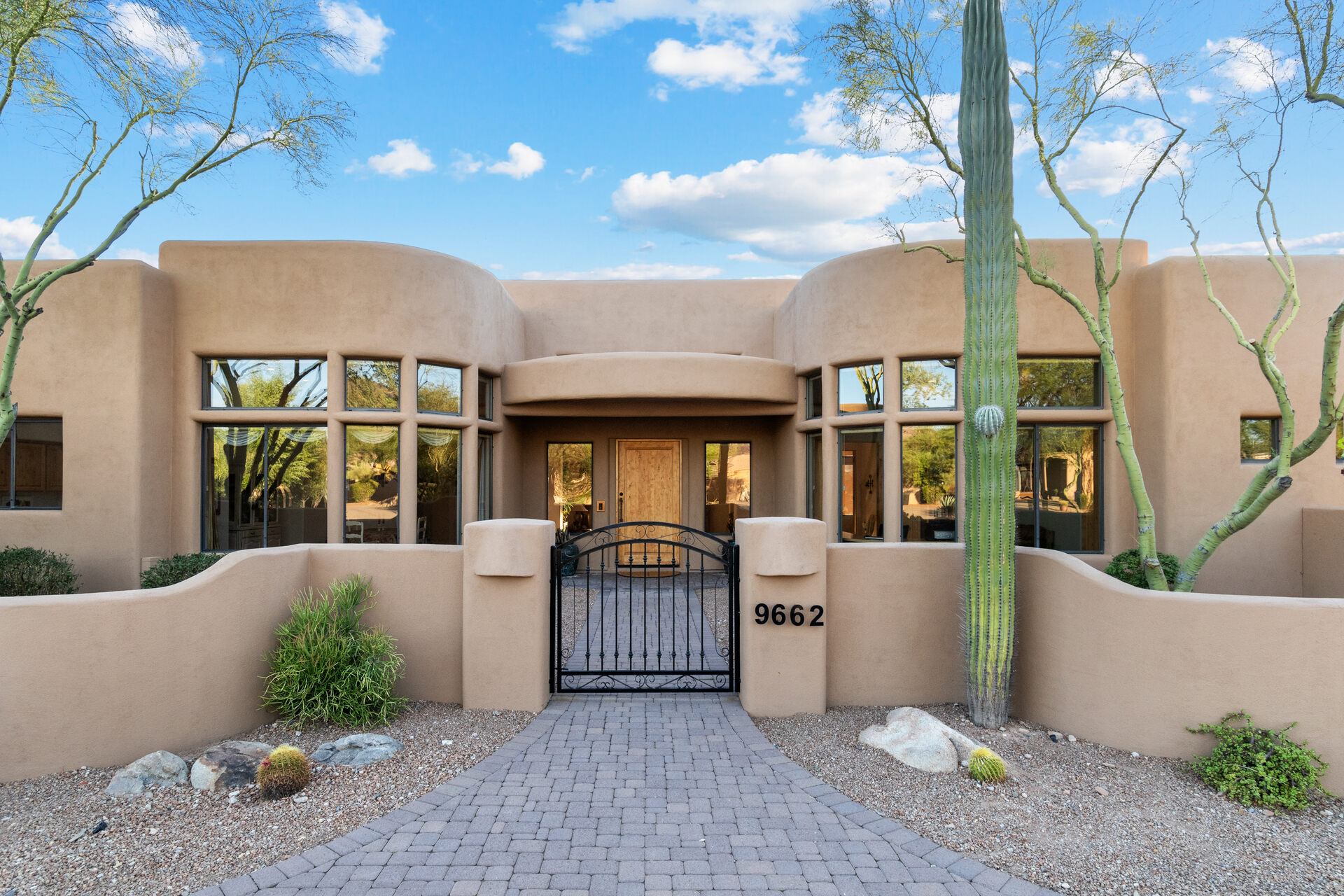 The image shows a modern Southwestern-style home with a smooth stucco exterior in a warm beige color. The entrance features a black wrought-iron gate leading to a wooden front door, framed by large windows that reflect the desert landscape. A tall saguaro cactus and desert plants adorn the front yard, which is covered in gravel with a paved stone pathway. The blue sky with scattered clouds adds to the bright and inviting atmosphere of the home.