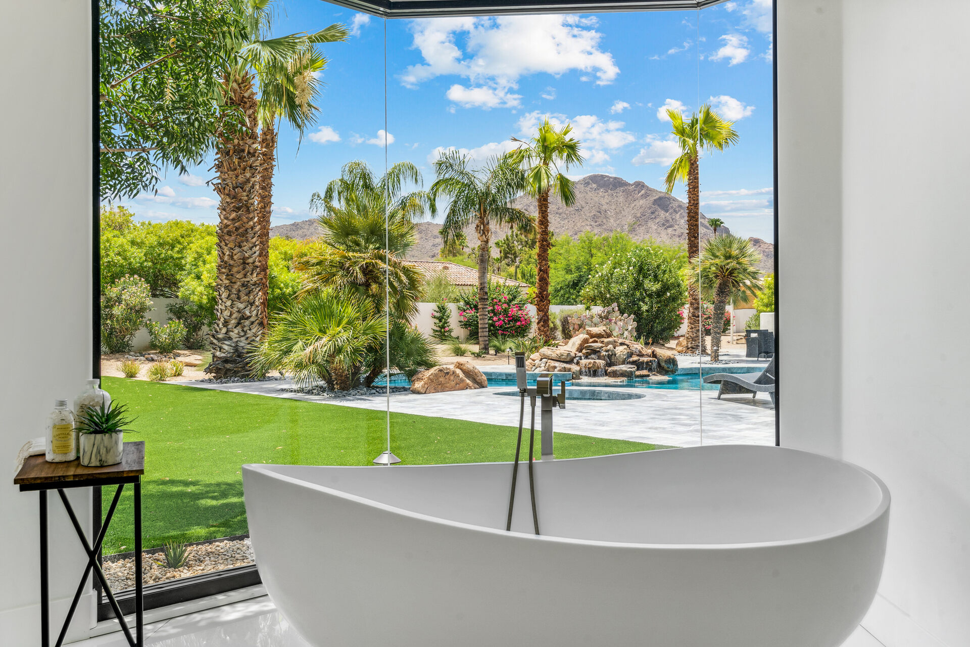 The image shows a luxurious bathroom with a modern freestanding bathtub positioned near a floor-to-ceiling glass window that overlooks a stunning backyard. The view includes lush greenery, palm trees, a pristine pool with a waterfall feature, and a backdrop of desert mountains under a bright blue sky. A small side table next to the tub holds bath products and a plant, adding a minimalist touch to the serene and elegant space. The natural light pouring in enhances the tranquil and spa-like atmosphere.