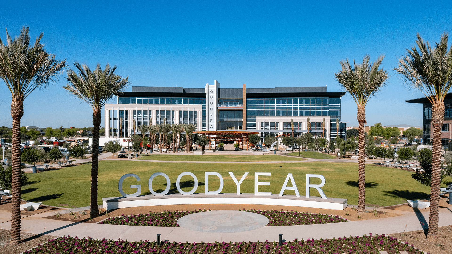 A wide shot of the Goodyear Square District in Goodyear, Arizona, featuring a large modern building with glass windows and the word 'Goodyear' displayed vertically on a central pillar. The foreground showcases a landscaped area with palm trees, green grass, and a flower bed, along with a large 'GOODYEAR' sign in bold letters. The sunny, blue sky adds to the vibrant scene.