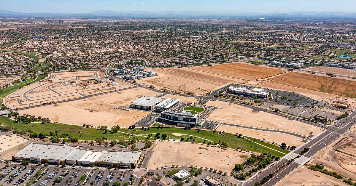 An aerial view of the new Goodyear Square District in Goodyear, Arizona, showcasing ongoing development with several large modern buildings, parking areas, and surrounding undeveloped land. The district is surrounded by residential neighborhoods, roads, and commercial spaces, with visible construction and infrastructure work in progress. The expansive view highlights the potential for future growth in the area.