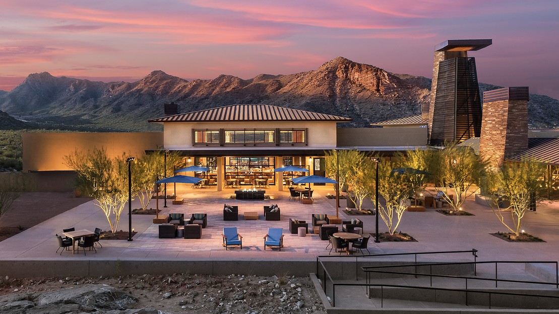 This image showcases the stunning Victory Clubhouse at Verrado. The clubhouse features a modern design with expansive outdoor seating areas, illuminated by soft lighting. Blue umbrellas provide shade over some seating areas, and a firepit adds warmth to the relaxing atmosphere. Behind the clubhouse, the rugged mountain landscape rises dramatically, bathed in the glow of a beautiful sunset with vibrant hues of pink and orange in the sky. The surrounding desert plants, including tall trees with subtle lighting, blend the natural beauty of the desert with the luxury of the clubhouse.