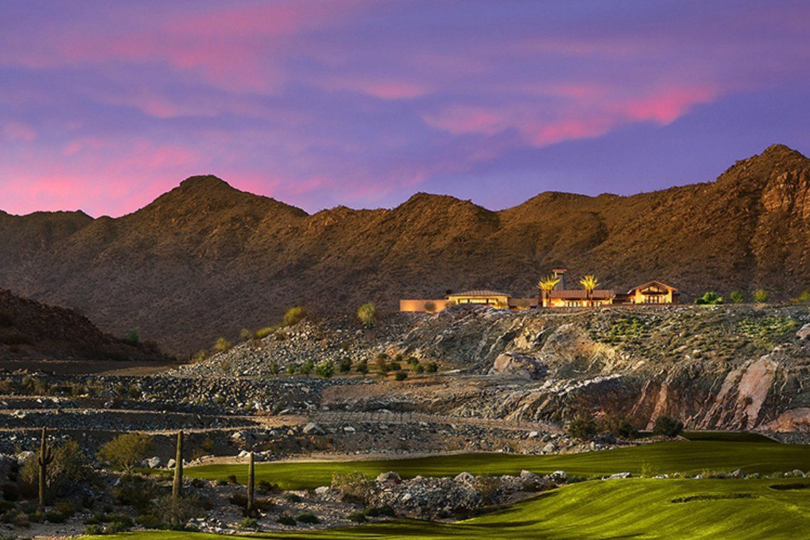 This image depicts a scenic view of Victory at Verrado, a beautiful community nestled against desert mountains with a rugged landscape in the foreground. The sunset sky casts a warm pink and purple hue over the mountains, contrasting with the green golf course and sparse desert vegetation below. A modern architectural structure is visible in the distance, blending into the natural surroundings, symbolizing the luxurious and tranquil lifestyle of the community.