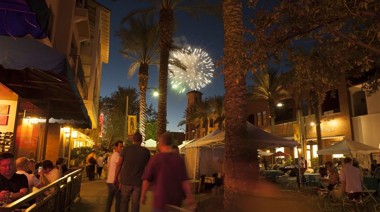 This image captures the vibrant atmosphere of downtown Verrado during an evening event. Palm trees line the lively streets, with people strolling through the area while enjoying the festivities. Outdoor dining spaces are filled with patrons, and small market stalls add to the community feel. In the background, a fireworks display lights up the night sky, casting a magical glow over the scene. The soft lighting from nearby buildings creates a warm, inviting ambiance, reflecting the charming and social atmosphere of Verrado's downtown events.