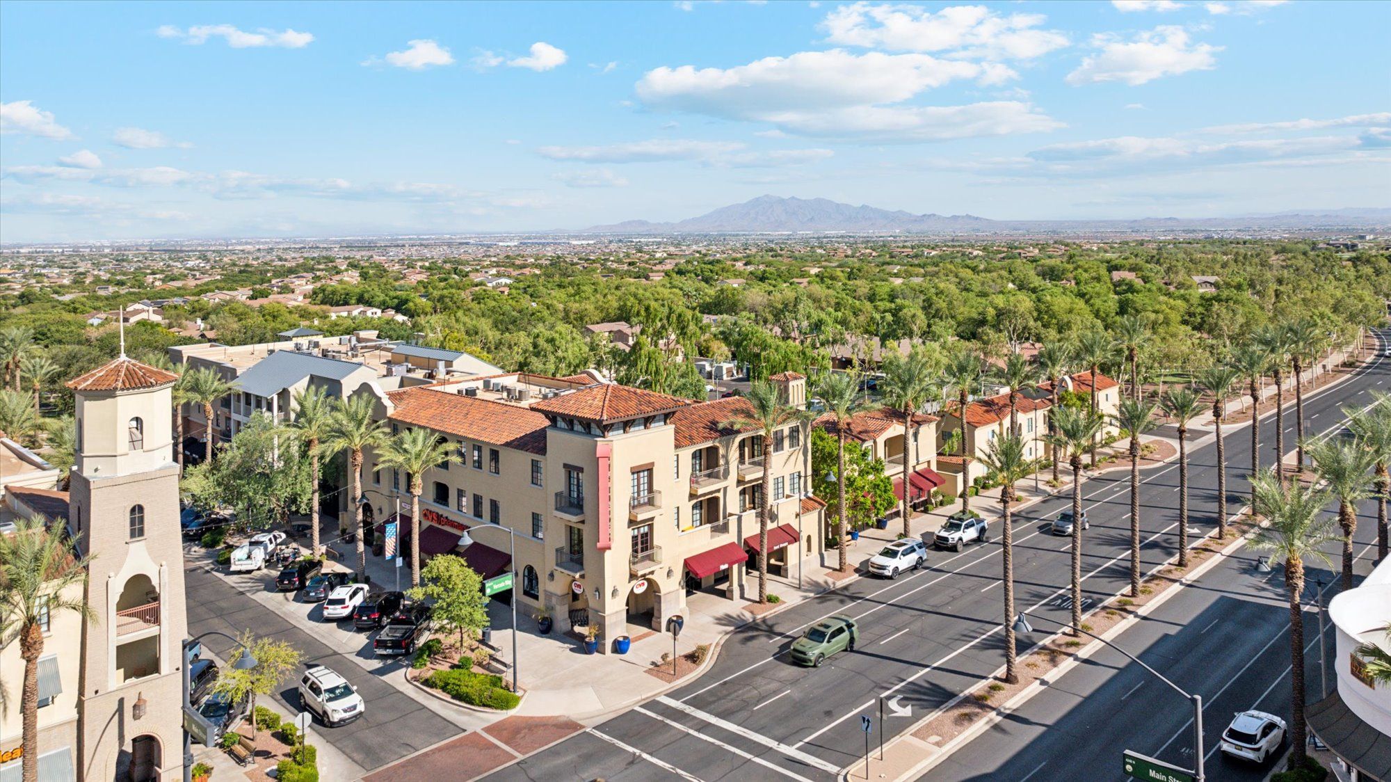 The image shows an aerial view of the Verrado community in Buckeye, Arizona. The scene captures a charming downtown area with Spanish-style architecture featuring red-tiled roofs and palm trees lining the streets. A central tower stands out as a key architectural feature. In the distance, lush greenery surrounds residential homes, creating a scenic contrast between the urban and natural landscape. The streets are well-maintained, and you can see a few cars and parked vehicles, giving the impression of a quiet yet active community. The backdrop includes a view of the distant mountains under a clear, bright sky.