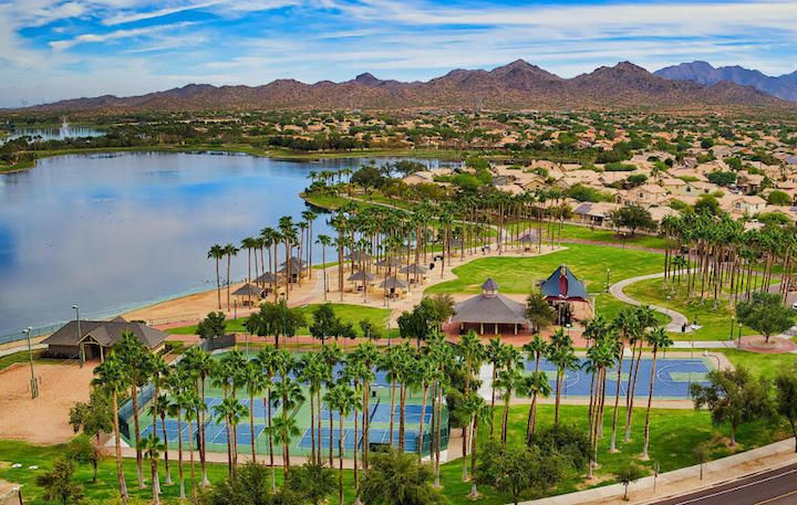 The image shows an aerial view of the Estrella community in Goodyear, Arizona, featuring a scenic lake surrounded by palm trees and recreational facilities. In the foreground, tennis courts and a small park area with pavilions are visible, offering a glimpse of the community’s outdoor amenities. The expansive lake is framed by lush greenery and walking paths, leading to residential neighborhoods with homes nestled in the distance. Beyond the community, the Estrella Mountains rise majestically, creating a picturesque backdrop. The bright blue sky with a few scattered clouds adds to the peaceful and inviting atmosphere of the Estrella community.