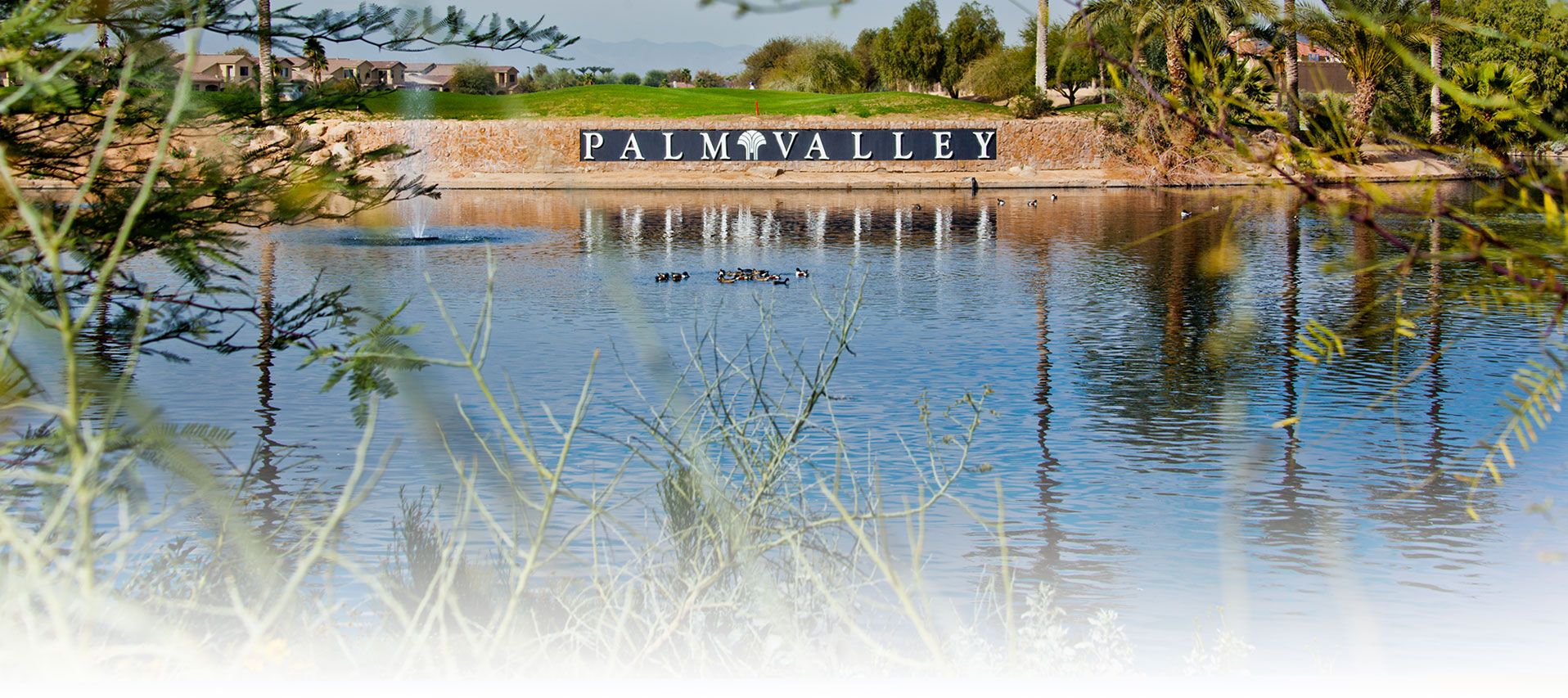 The image shows a serene view of a large pond or small lake in Palm Valley, a community located in Goodyear, Arizona. Ducks are swimming in the water, and a fountain is visible in the background. The focal point is a large stone sign that reads "PALM VALLEY," situated on the far side of the water. In the distance, you can see well-maintained greenery, palm trees, and residential homes, contributing to the peaceful, suburban feel of the Palm Valley community. The foreground is framed by desert foliage, enhancing the natural beauty of this picturesque area in Goodyear, Arizona.