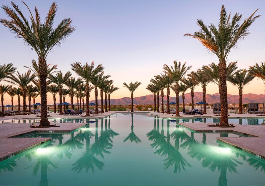 The image shows a luxurious, resort-style pool at Sterling Grove in Surprise, Arizona. The pool is lined with tall, perfectly spaced palm trees, creating a symmetrical and inviting scene. The water reflects the trees and the soft lighting as the sun sets behind the mountains in the background, casting a warm glow over the landscape. The sky transitions from light to darker hues, enhancing the serene and relaxing atmosphere. Lounge chairs and cabanas are placed around the pool, providing a comfortable space for relaxation. The distant mountains and desert landscape add a stunning natural backdrop to the peaceful setting.