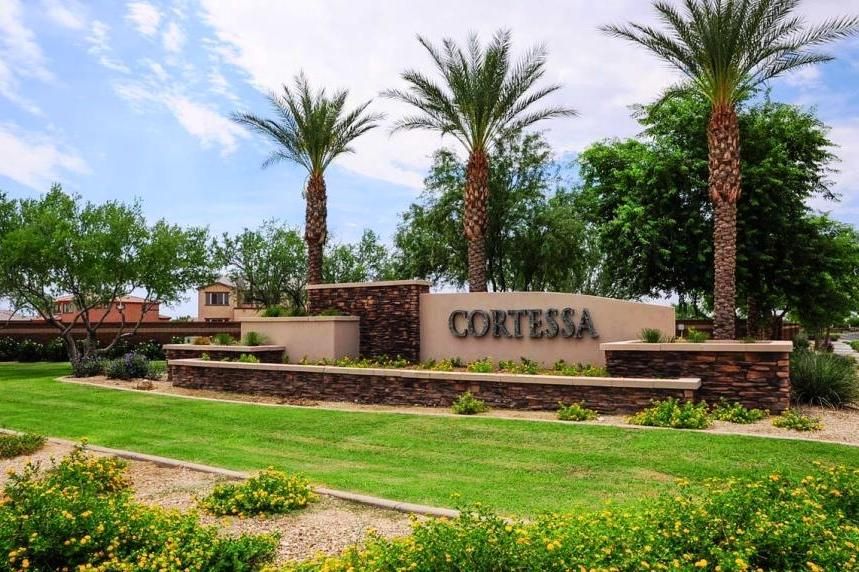 The image shows the entrance sign to the Cortessa community in Waddell, Arizona. The sign, which reads "Cortessa," is mounted on a stone and stucco structure surrounded by neatly landscaped greenery and colorful flowers. Tall palm trees and lush trees frame the scene, adding to the well-maintained and inviting atmosphere. The bright green grass and desert-friendly plants enhance the beauty of the entrance, while the blue sky and scattered clouds provide a serene backdrop, creating a welcoming first impression of the community.