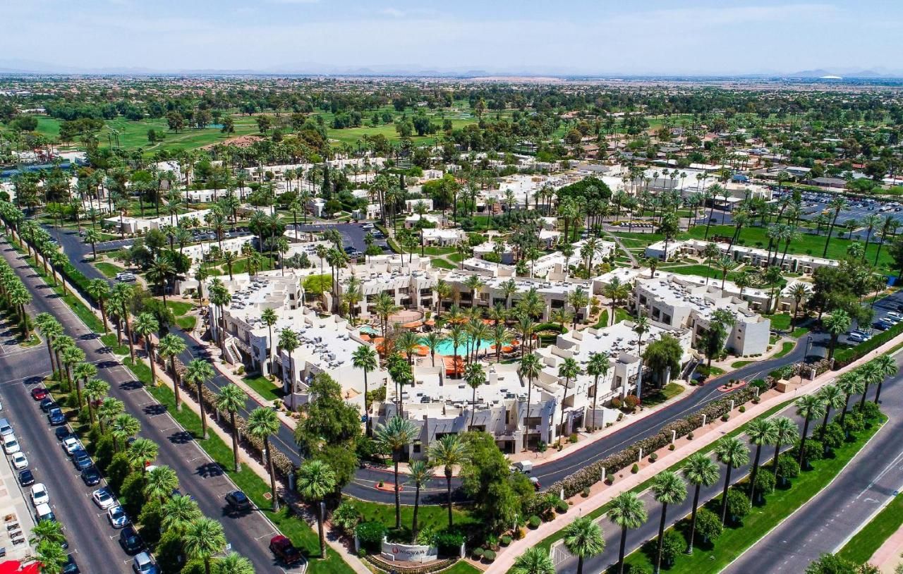 The image shows an aerial view of the Wigwam, a luxury resort located near the Litchfield Greens community in Litchfield Park, Arizona. The resort features distinct white adobe-style buildings, lush landscaping, and palm-lined streets. At the center, there is a large pool surrounded by palm trees and vibrant orange umbrellas, providing a focal point of relaxation. In the background, the Wigwam's expansive golf courses stretch out into the distance, bordered by green fields and suburban homes. The surrounding area captures the essence of resort-style living with wide, well-maintained roads and scenic views of the desert landscape.