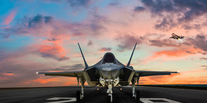 This image shows a close-up of an F-35 fighter jet positioned on the runway at Luke Air Force Base in Arizona. The jet is ready for takeoff at sunset, with a second F-35 flying in the background. The vibrant colors of the sky, transitioning from orange to pink and blue, enhance the dramatic setting, highlighting the sleek design of the fighter jets. The open runway and distant horizon emphasize the vast, open desert landscape characteristic of the base's location.