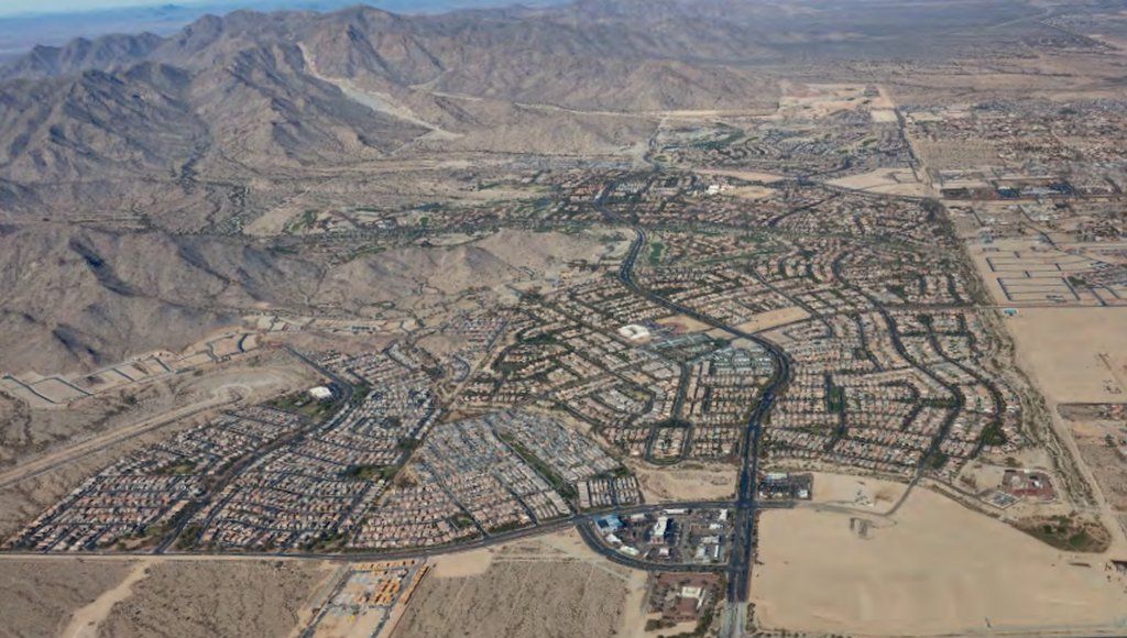 This image provides an aerial view of Verrado, one of the most popular neighborhoods among those stationed at Luke Air Force Base. The community is nestled at the base of the White Tank Mountains in Buckeye, Arizona, with a sprawling layout of residential homes and carefully planned streets. The image showcases the desert landscape surrounding the area, with the rugged mountain terrain offering a scenic backdrop. Verrado’s master-planned design is evident, with green spaces, parks, and schools integrated throughout the community, making it a desirable location for military families. The vast open desert contrasts with the orderly, vibrant neighborhood below.