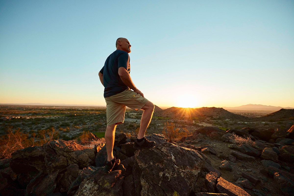 A scenic image of a resident hiking in Verrado, Buckeye, AZ, captured at sunrise. The hiker stands atop rugged rocks, gazing out over the vast desert landscape, with the warm glow of the sun rising behind the distant mountains. This image beautifully showcases the outdoor lifestyle and natural beauty that Verrado offers, highlighting its appeal for residents who enjoy hiking and exploring the Arizona desert. The peaceful and expansive surroundings create a sense of adventure and tranquility.