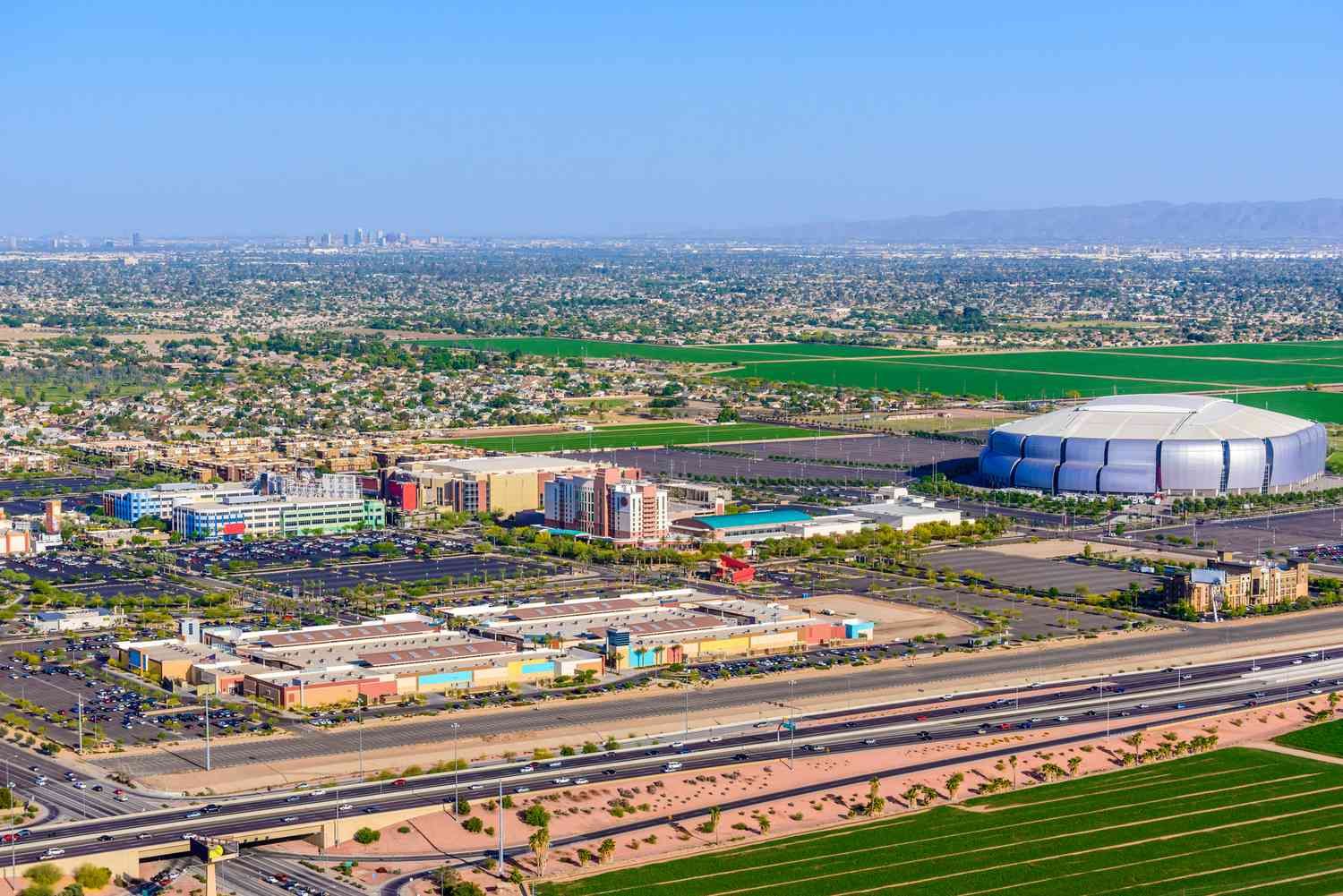 An aerial view of the Westgate Entertainment District in Glendale, Arizona, featuring the iconic State Farm Stadium, home of the Arizona Cardinals. The stadium, known for hosting major events like the Super Bowl, the Final Four, and various concerts, stands out in the landscape. Surrounding the stadium are entertainment venues, hotels, and shopping areas that make Westgate a bustling hub for sports fans and visitors. The expansive view showcases the district's accessibility, located near major freeways, with the Phoenix skyline visible in the distance.