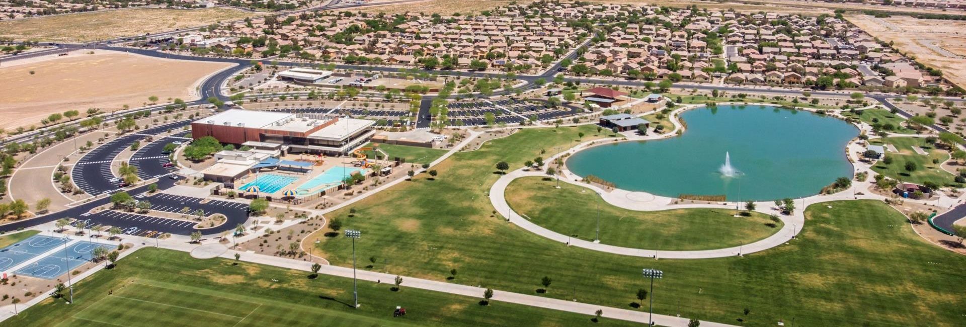 Aerial view of Maricopa, Arizona, showcasing a large community park with a lake, sports courts, swimming pools, and nearby residential areas. The image highlights the well-planned infrastructure and recreational amenities that contribute to Maricopa's appeal as a growing suburb.