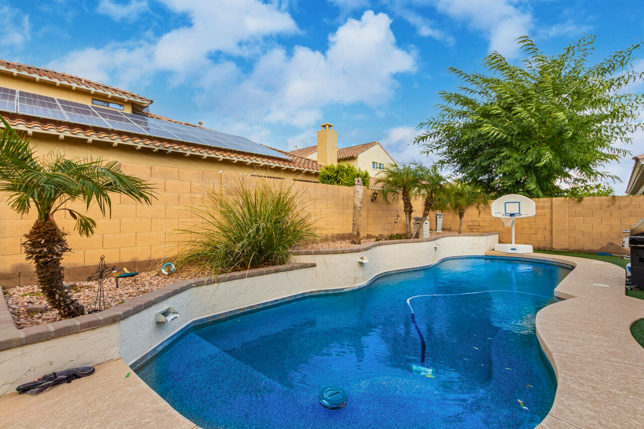 Beautiful backyard with a sparkling pool in a Palm Valley home, Goodyear, AZ. The space features a curved swimming pool, palm trees, and low-maintenance landscaping, offering an ideal outdoor area for relaxation or entertainment. Solar panels are visible on the roof, adding energy efficiency to this desirable property in one of Goodyear’s premier neighborhoods.