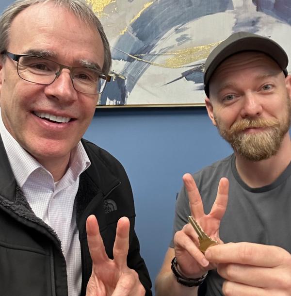 David stands next to Nick, both smiling and holding up two fingers. Nick has his house key held in front of the camera.