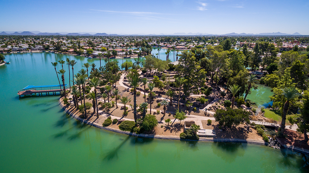 Scenic aerial view of Sun City, Arizona, showcasing serene turquoise lakes, lush greenery, palm trees, and surrounding homes under a clear blue sky, highlighting the tranquil and picturesque lifestyle of this retirement-friendly community.