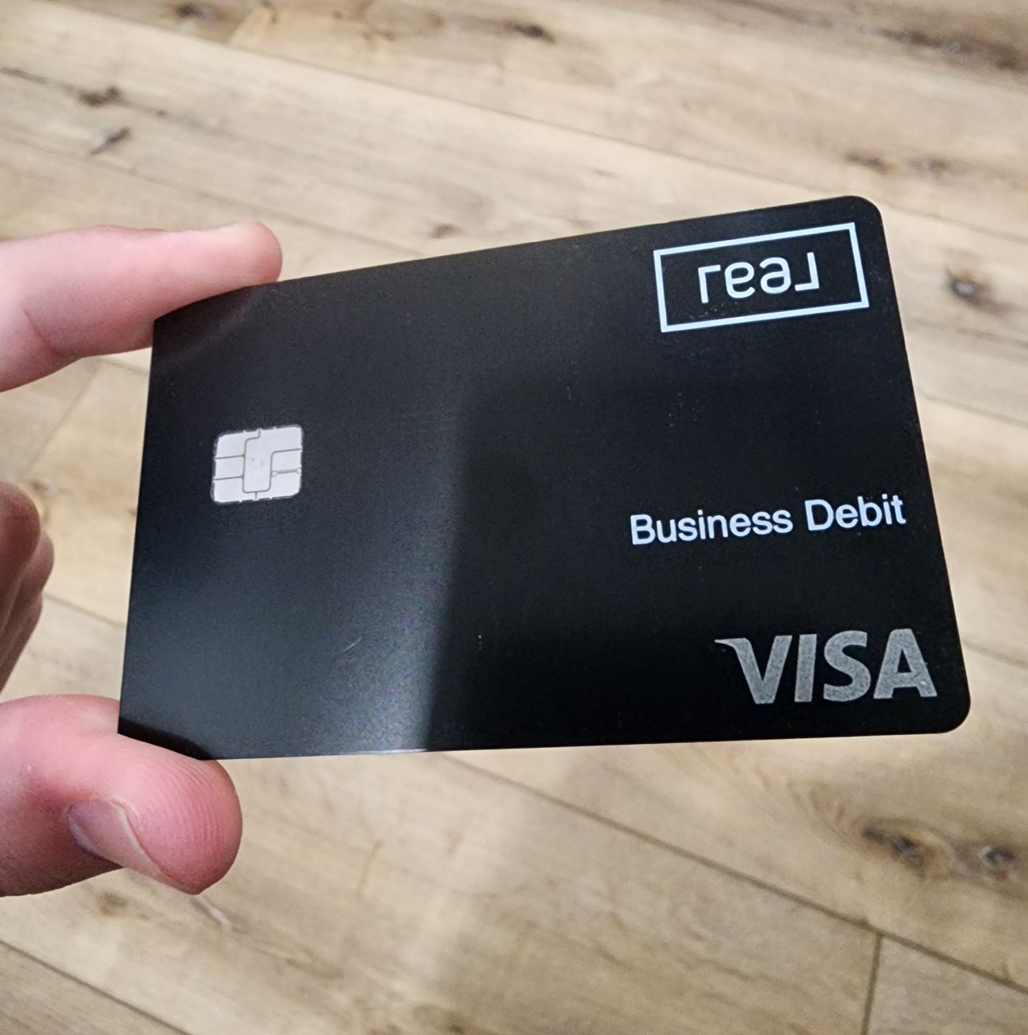 Real Broker Real Wallet Business Debit Card, featuring a sleek black design with Visa logo, held against a wooden floor background, showcasing a modern financial solution for real estate professionals.