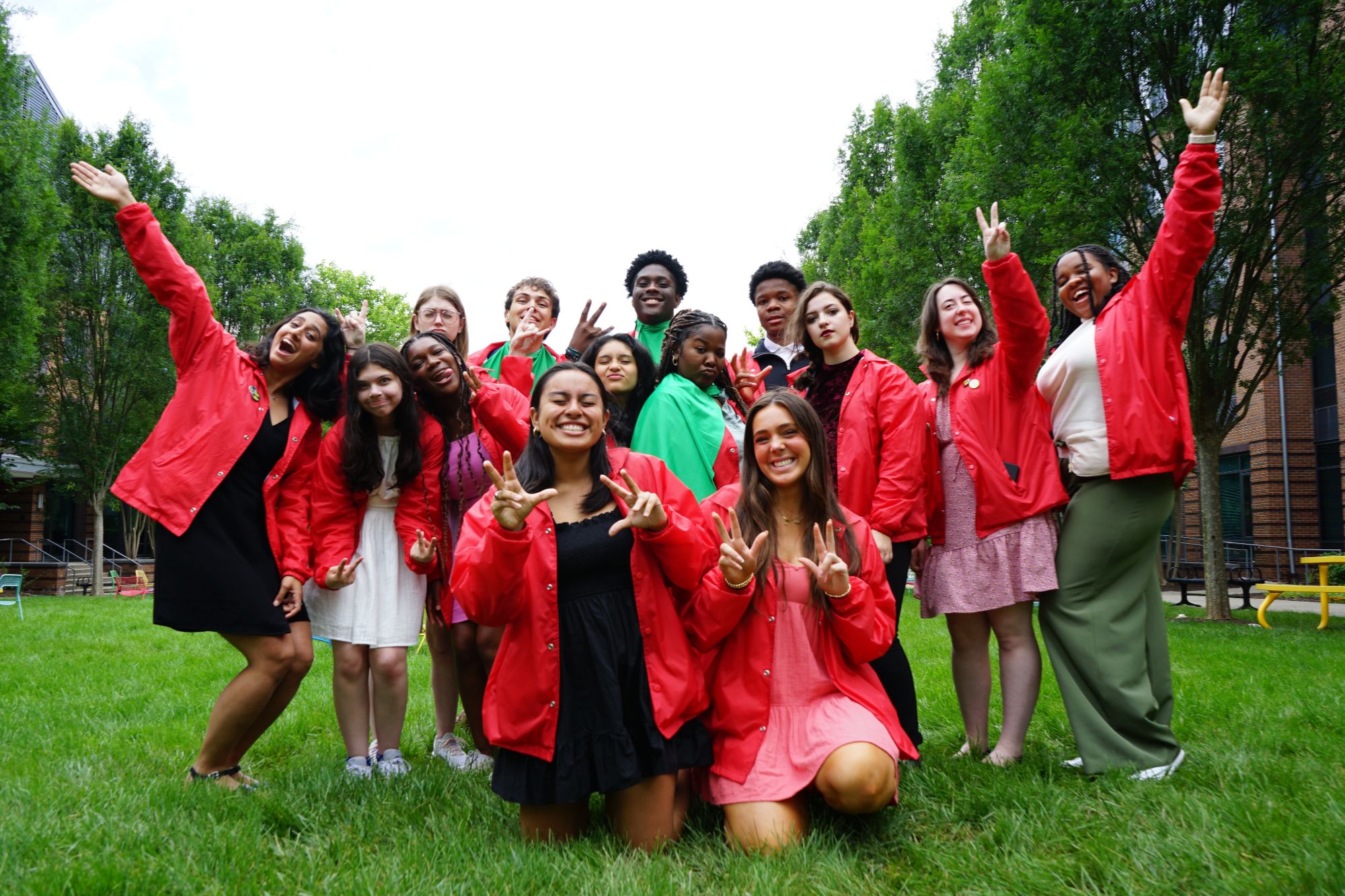 A lively group of HOBY Maryland participants wearing red jackets, smiling and posing energetically on a grassy lawn with trees and buildings in the background.