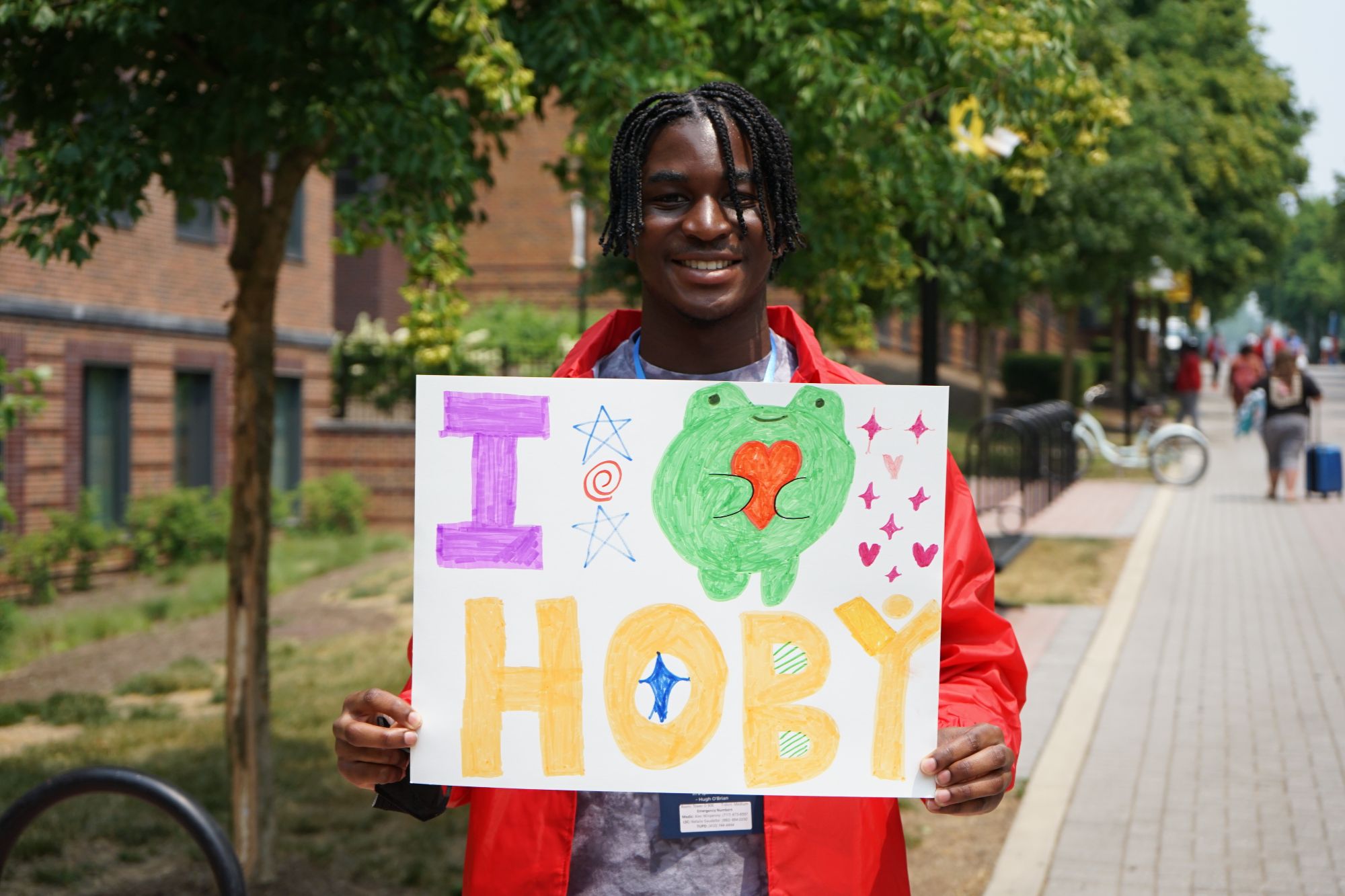 A smiling HOBY Maryland participant holding a colorful handmade sign that reads 'I ❤️ HOBY' with playful drawings, standing outdoors on a sunny day.