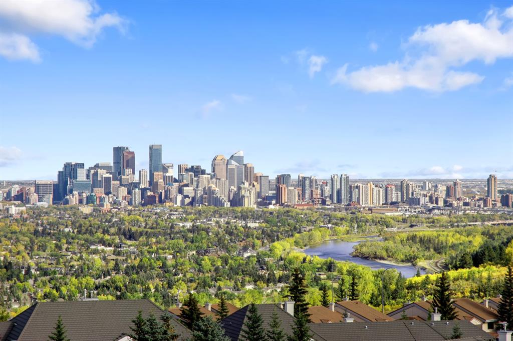 A stunning view of Calgary's downtown skyline with modern skyscrapers, surrounded by lush green trees and the winding Bow River. The foreground showcases rooftops of residential homes, adding depth to the image under a bright blue sky with scattered clouds.