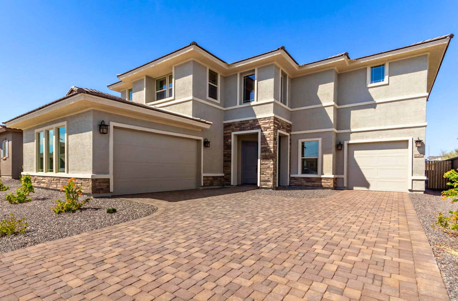 Modern two-story new construction home in Litchfield Park, AZ, featuring a spacious three-car garage, stone-accented exterior, and paved driveway under a clear blue sky. The contemporary design showcases large energy-efficient windows and clean architectural lines, ideal for luxury living.