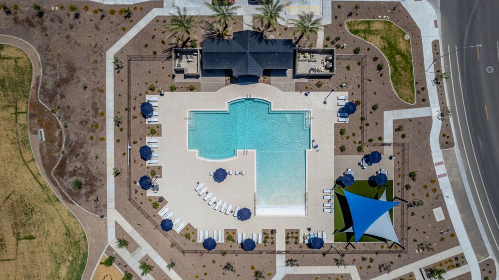 Aerial view of a resort-style community pool in Litchfield Park, AZ, featuring a large T-shaped swimming pool surrounded by lounge chairs, shaded seating areas with blue umbrellas, and a covered pavilion. Modern landscaping and walkways highlight the upscale amenities of this master-planned community.