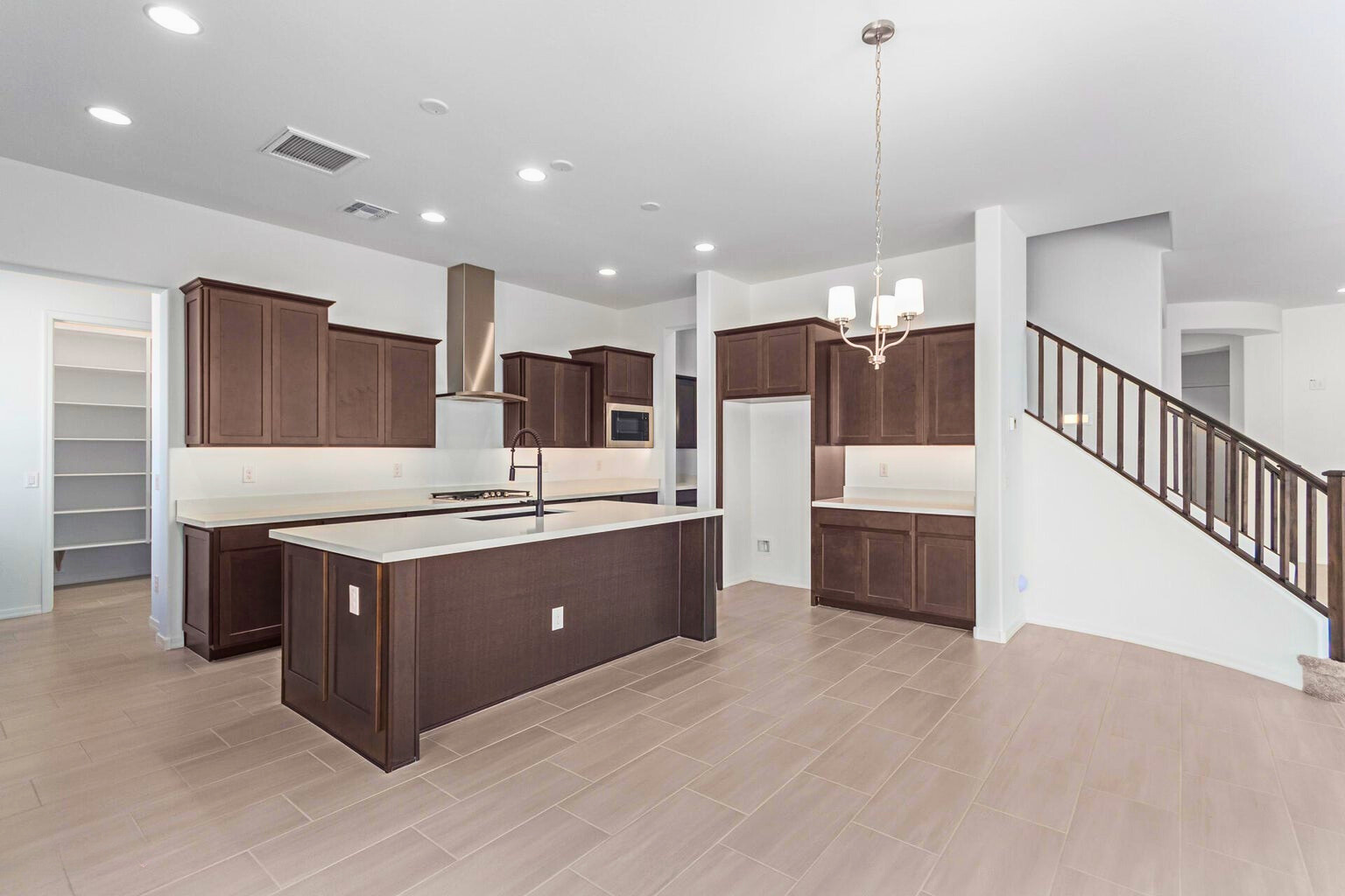 Spacious modern kitchen in a new build home in Litchfield Park, AZ, featuring dark wood cabinetry, a large island with quartz countertops, stainless steel range hood, walk-in pantry, and elegant staircase leading to the second level. Bright open-concept layout with recessed lighting and natural light.