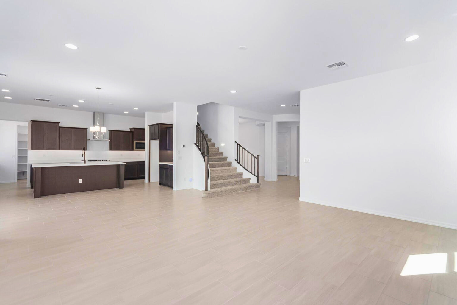 Spacious open-concept living area in a new build home in Litchfield Park, AZ, featuring modern dark wood cabinetry in the kitchen, a large island, walk-in pantry, and a central staircase with elegant railings leading to the second floor. Natural light enhances the bright, airy layout.