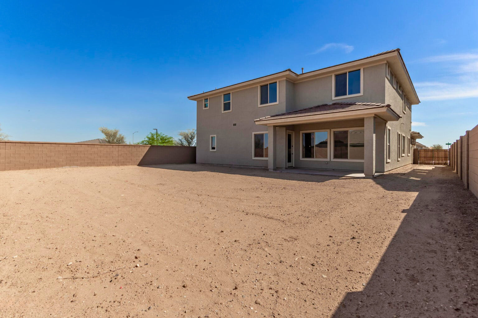 Spacious backyard of a new build home in Litchfield Park, AZ, featuring a two-story modern design with covered patio space and large windows. The expansive lot offers a blank canvas for landscaping or outdoor living in a private, walled setting under a clear blue sky.