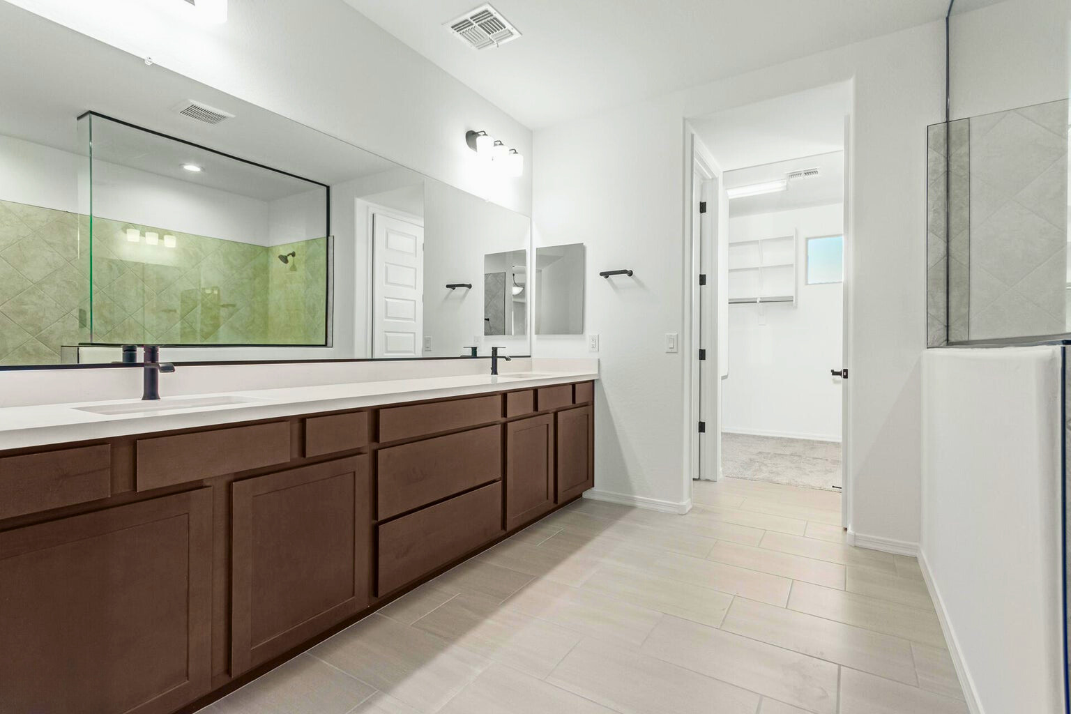 Modern primary bathroom in a new build home in Litchfield Park, AZ, featuring dual sinks with dark wood cabinetry, a large mirror, a glass-enclosed walk-in shower, and access to a spacious walk-in closet. Bright, clean design with elegant finishes and neutral tile flooring.