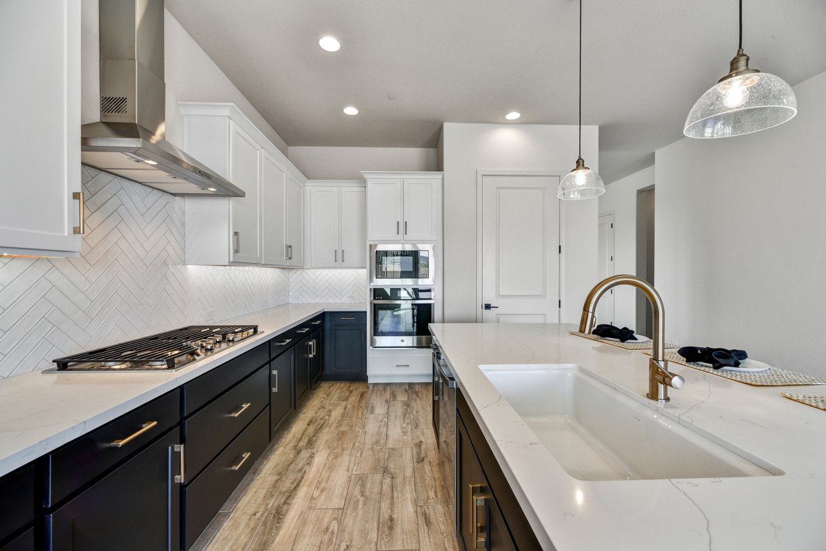 Elegant kitchen in a new construction home in Peoria, Arizona, showcasing a spacious quartz island with seating, stainless steel appliances, custom cabinetry, and a designer herringbone tile backsplash. The open layout features pendant lighting and bright natural light, making it an ideal space for cooking, dining, and entertaining.