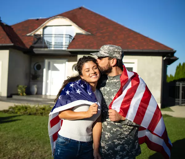 portrait-soldier-uniform-kissing-his-wife-holding-american-flag-front-their-house